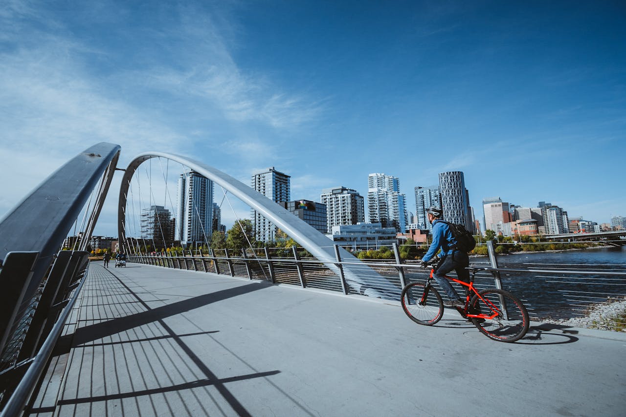 A cyclist rides across a modern bridge with a city skyline in the background, under a clear blue sky.
